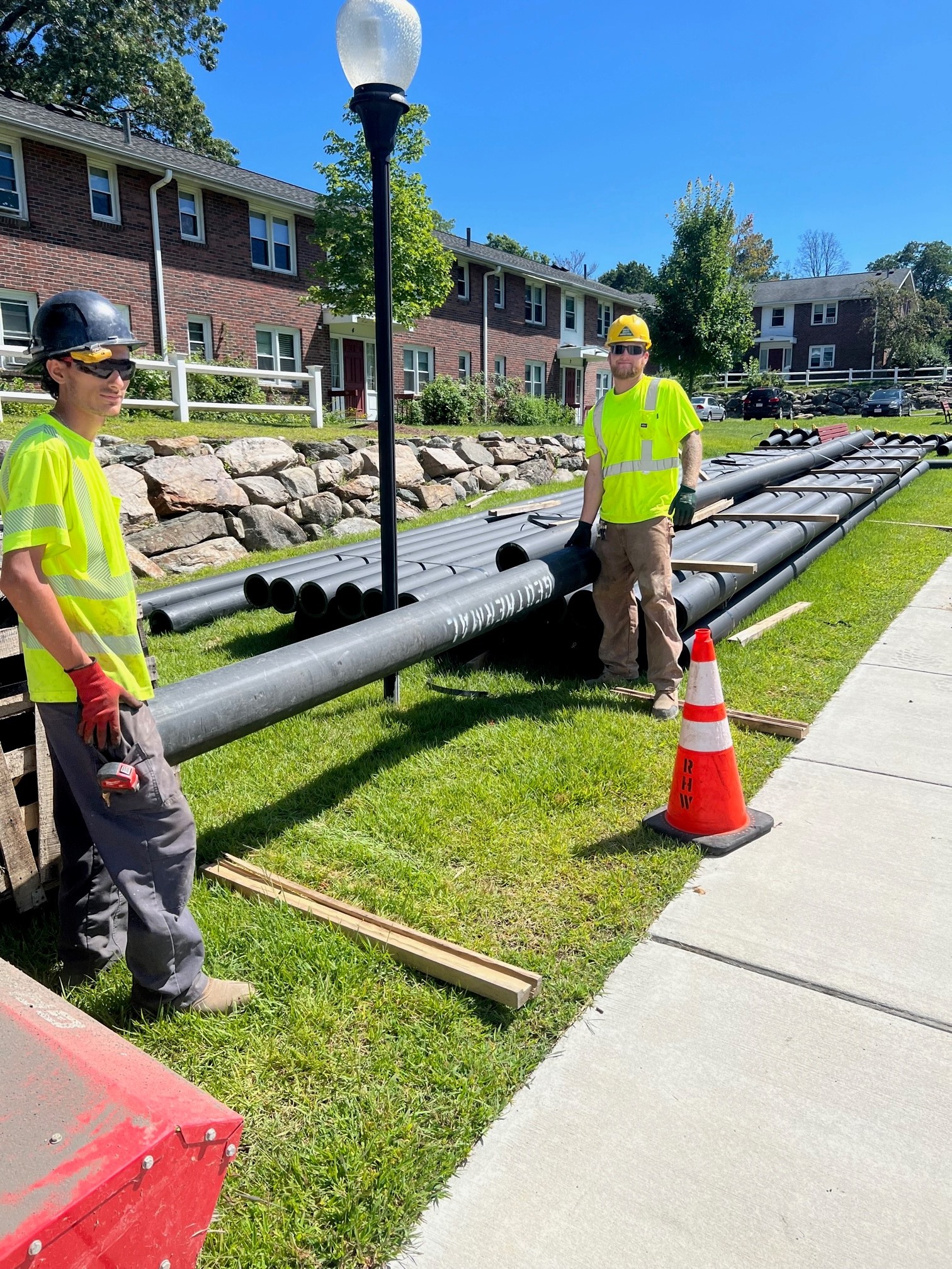 Two construction workers in safety gear standing next to rows of black HDPE ambient-loop pipes on the grass, in front of a brick apartment complex.